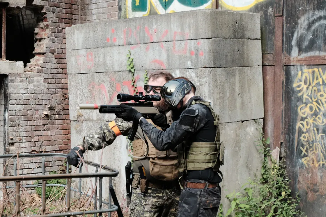 Two airsoft players behind cover by a low wall, one aiming a marksman replica, in an urban setting.