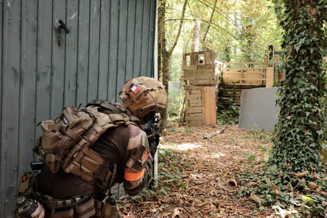 Airsoft player in gear and helmet, replica shouldered, holding cover along a wall on a woodland field.